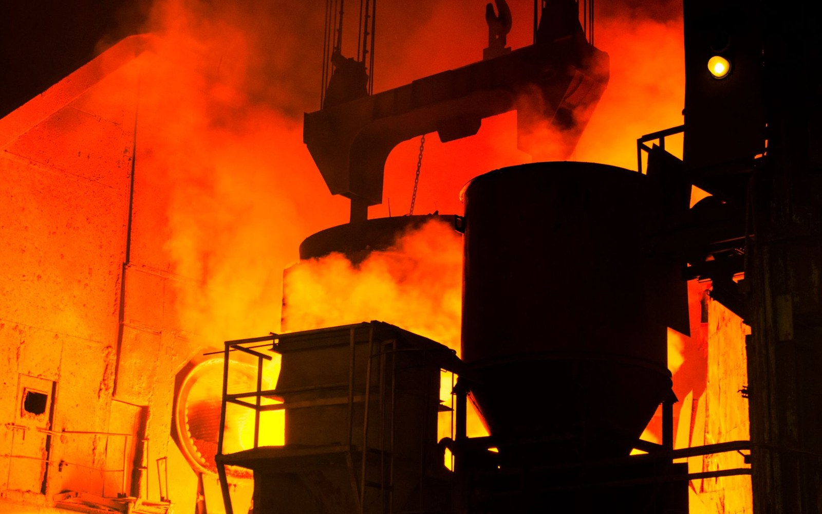 Molten metal glows orange inside a steel mill furnace with heavy smoke and machinery silhouetted