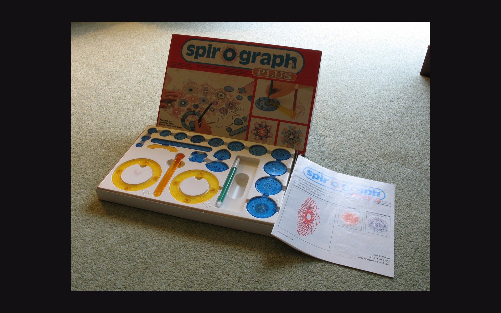 An open Spirograph Plus box showing colorful gears, wheels and an instruction sheet on carpet