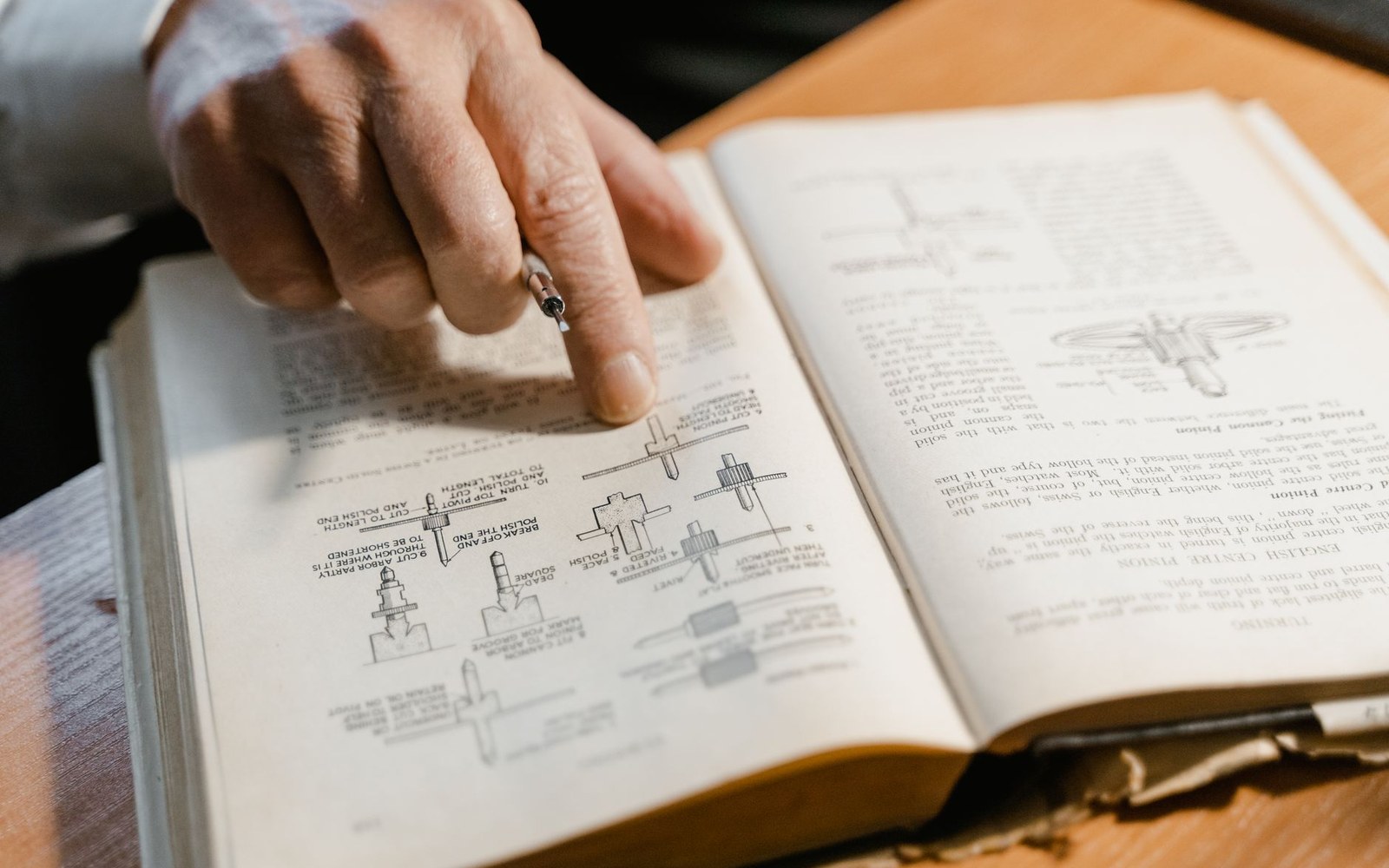 A hand holding a pencil points to mechanical diagrams in an open instructional book
