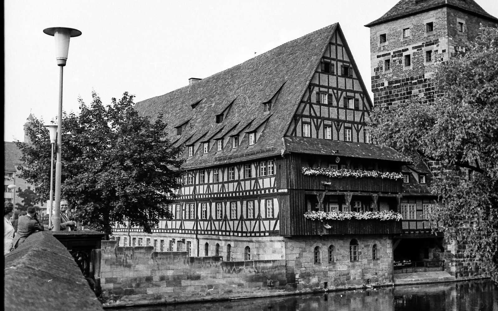 Half-timbered building with a steep gabled roof stands beside a river and stone tower